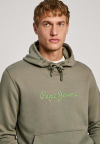 Sudadera verde oliva con cordón, que cuenta con un bolsillo frontal tipo canguro y un logo bordado de "Pepe Jeans" en verde claro. Tela suave y texturizada.