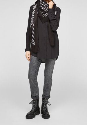 Personne portant une tunique noire, une écharpe rayée noir et blanc, un jean gris retroussé et des bottes noires à lacets, debout sur un fond gris.