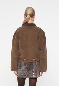 Bruine corduroy jas met een opvallende kraag en schoudernaden, voorzien van ronde knopen en een relaxte pasvorm, gecombineerd met een paillettenrok.