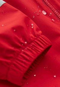 Manche de veste imperméable rouge avec poignet élastique et gouttes d'eau perlées sur le tissu près d'une fermeture éclair.