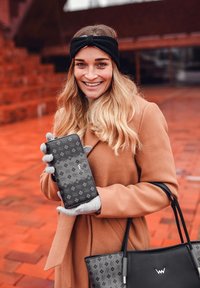Black patterned wallet with a smooth texture, held by a person in a tan coat and gray gloves, with a black handbag visible nearby.