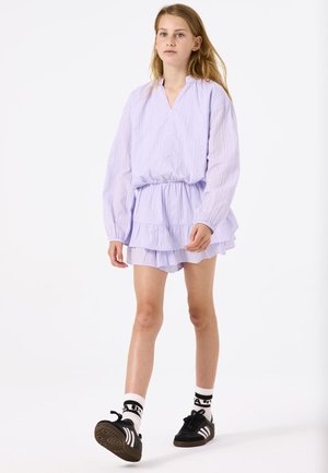 Robe lilas clair, texturée, avec des manches longues, un décolleté en V et une jupe à étages. Portée avec des chaussures noires et des chaussettes blanches avec des accents noirs.