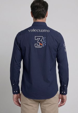 Hombre de espaldas con camisa azul marino abotonada, con "valecuatro" y el número 3 con el texto "ESP" bordados en la espalda y la manga.