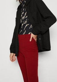 Zwarte gestructureerde jas met een kraag en knoopsluiting, gecombineerd met een gemêleerd bodysuit en rode slimfit broek.