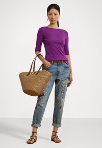 Paarse getailleerde top, versleten jeans met verfspetters, bruine geweven handtas en bruine sandalen met bandjes, staand op een neutrale achtergrond.