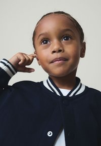 Otrok s temnimi lasmi v mornarsko modri jakni si dotika uho, pogleda pa rahlo navzgor in v stran na enostavni ozadju.