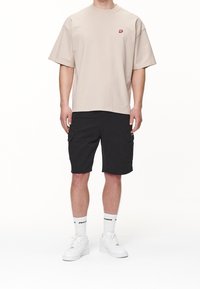 Beige oversize tričko s malým červeným logem, spárované s černými cargo šortkami s bočními kapsami, bílými teniskami a bílými ponožkami.