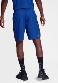 Pantalones cortos atléticos azules con rayas en forma de chevron negras a los lados, cinturilla elástica y ajuste relajado, combinados con calcetas negras hasta la rodilla y zapatillas deportivas.