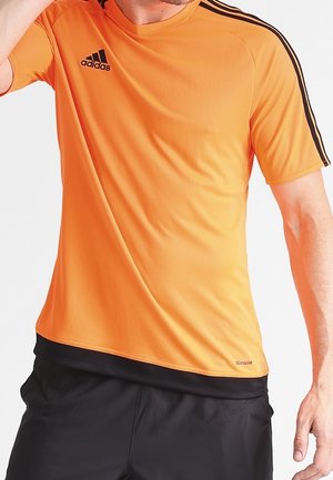 Mężczyzna noszący pomarańczową sportową koszulkę Adidas z krótkim rękawem, z czarnymi paskami na ramionach i czarnym logo Adidas, w połączeniu z czarnymi spodniami krótkimi.