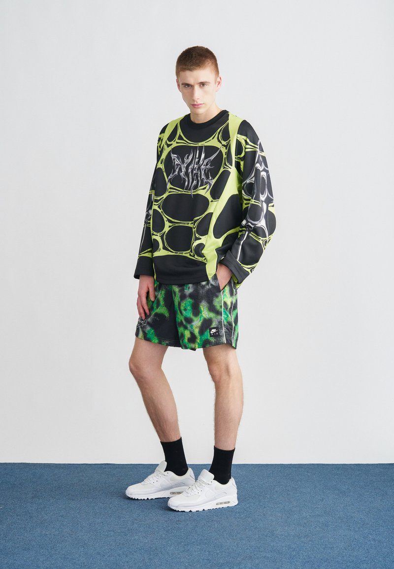 Black and neon green long-sleeve shirt with abstract patterns, paired with green and black patterned shorts and white sneakers.