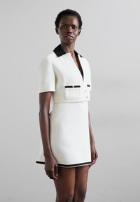 Weißes kurzes Kleid mit strukturierter Oberfläche, schwarzem Kragen und Akzenten. Mit kurzen Ärmeln, Knopfverschluss vorne und Taschen.