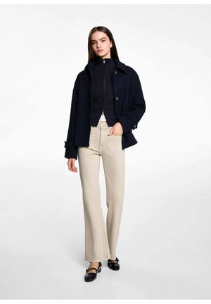Marineblaue Wollmischung Jacke, gerippter Strick-Zip-Up-Top, beigefarbene Schlagjeans und schwarze Lackflats. Das Modell steht vor einem neutralen Hintergrund.