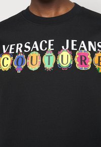 Svart crewneck-tröja med "VERSACE JEANS COUTURE" i fet vit text och färgglada ramdesigns, vilket skapar en livfull visuell kontrast.