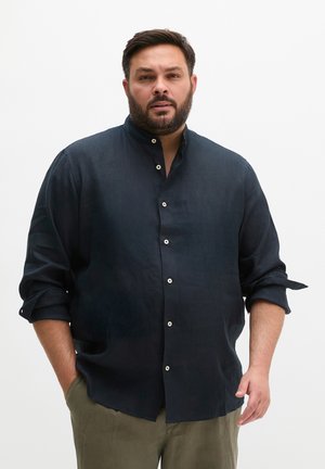 Mann mit Bart, der ein dunkles Hemd mit Knöpfen und olivfarbene Hose trägt, steht mit einer Hand in der Tasche vor einem schlichten Hintergrund.