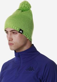 Berretto a maglia verde con pompon in cima, con un'etichetta nera con un logo. Indossato con una giacca blu navy con zip a metà.