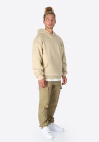 Beige Kapuzenpullover mit Fronttasche, weißem Lagenhemd, olivfarbenen Cargo-Hosen und weißen Sneakers. Einfaches Design mit minimalen Verzierungen.
