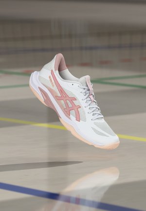 Witte sportschoen met roze accenten zwevend boven een gymvloer met gekleurde lijnen voor sportvelden.