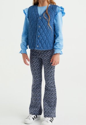 Blauw gewatteerd vest met vijf knopen, gecombineerd met een lichtblauw textuurshirt en patroonachtige donkerblauwe flared broeken. Witte sneakers maken de outfit compleet.