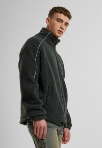Dark grey fleece jacket with high collar, zip closure, and white accent stitching. Features front pockets and a textured, warm fabric.