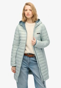 Lys blå lang quiltet frakke med hætte, lodret quiltet tekstur, lynlås lukning og logo på brystet, båret over en cremefarvet sweater og blå jeans.