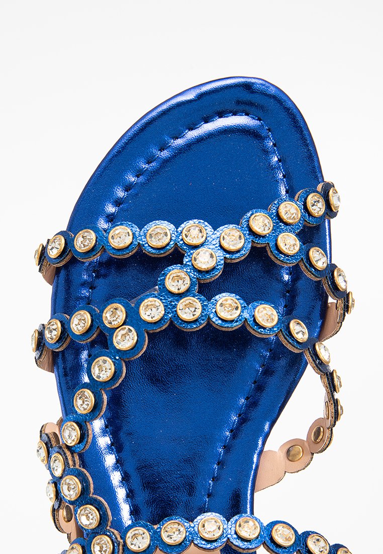 Sandali metallici blu con cinturini ondulati decorati con strass circolari. Parte superiore testurizzata e soletta liscia con fibbia laterale regolabile.