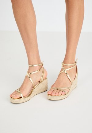 Sandales compensées dorées à brides avec une plateforme en jute tissé, au design élégant et à bout ouvert, mettant en valeur les pieds.