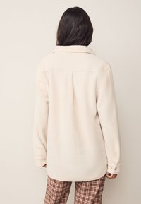 Giacca in pile color crema con collo con risvolto, polsini con bottoni a pressione e vestibilità rilassata, mostrata di schiena su uno sfondo neutro.