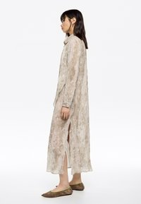 Vestido maxi de color beige claro y transparente, con un patrón de serpiente, aberturas en los laterales, mangas largas y puños abotonados; combinado con zapatos planos marrones con un acento verde.