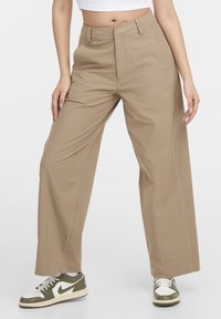 Personne portant un pantalon beige à jambes larges, un haut court blanc, et des baskets Nike blanches et vert olive, debout les jambes écartées.