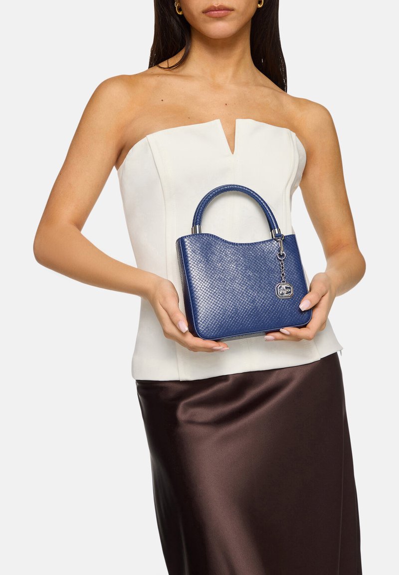 Blau strukturierte Handtasche mit einer festen Form und kurzem Griff, mit einem silbernen Charme-Detail. Getragen mit einem weißen Oberteil und einem braunen Rock.