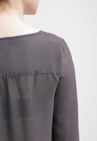 Grå, genomskinlig blus med en rundad halsringning, som har en samlad detalj i ryggen och långa, åtsittande ärmar. Slät struktur med ett lätt utseende.