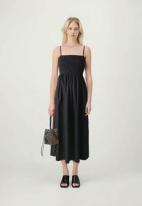 Kvinna iklädd en svart ärmlös midiklänning med smockad byst, håller en liten texturerad handväska och svarta öppna halkskor.