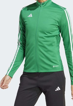 Persoon die een groen ritsjack met witte strepen op de mouwen draagt en een zwarte broek, beide met Adidas-logo's.