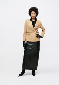 Μπεζ διπλόμπουτο σακάκι με λεπτομέρεια χρυσής αλυσίδας, σε συνδυασμό με μαύρη δερμάτινη μακριά φούστα και μαύρα γάντια. Το μοντέλο ποζάρει σε ουδέτερο φόντο.