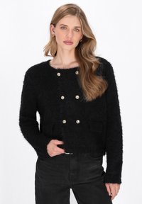 Zwarte, fluffy cropped cardigan met dubbelrijige gouden knopen, ronde halslijn en voorzakken, gedragen over donkere jeans.