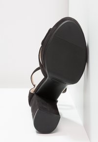 Sandale à talons hauts en daim noir avec une semelle en caoutchouc noir texturé, dotée de fines brides et d'un design de talon fermé.