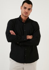 Camisa negra de manga larga con cuello abotonado, tejido texturizado y botones en la parte frontal. Presenta un dobladillo curvado y un diseño ajustado.