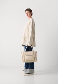 Persoon met kort haar, gekleed in een oversized beige trui, wijde manchet jeans, zwart en beige schoenen, en met een grote beige handtas tegen een witte achtergrond.