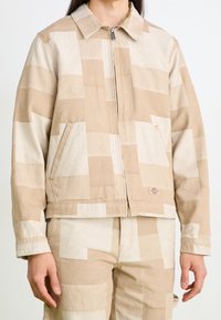 Beige vzorovaná bunda vyrobená z bavlny, s uzávěrem na zip, dvěma bočními kapsami a límcem, s čtvercovým blokovým designem v různých odstínech.