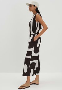 Zwart-wit gestippelde jumpsuit met mouwloos ontwerp, cinched taille en wijde pijpen. Model draagt een pet en platte sandalen.