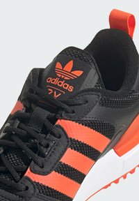 Scarpa sportiva nera Adidas con strisce arancioni, tomaia in rete, suola testurizzata e logo a contrasto sulla linguetta. Inclusi lacci e linguetta di estrazione.