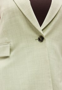 Εκρού σακάκι με ραφές προσαρμοσμένες στο σώμα, κλειστό με κουμπιά πάνω από σκούρο καφέ τοπ, διαθέτει γιακά με εγκοπή και πλαϊνή τσέπη με καπάκι στην αριστερή πλευρά.