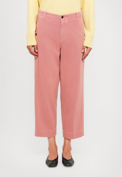 Persoon in roze wijde cropped broek, gele lange mouwen top en zwarte lage hakken, staande tegen een effen achtergrond.