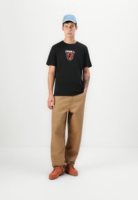 Sort t-shirt med rødt O'Neill-logo, beige løse bukser og orange sko. Blå hat fuldender outfittet. Hvid baggrund.