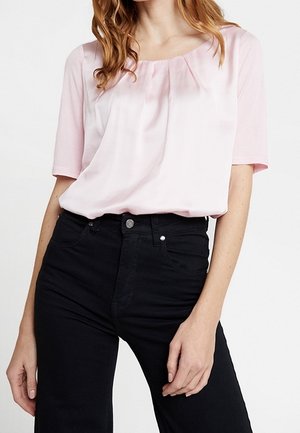 Kobieta ubrana w jasnoróżową bluzkę z krótkim rękawem i plisami, w połączeniu z czarnymi jeansami z wysokim stanem na białym tle.