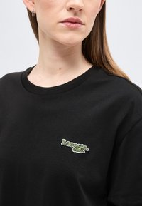 Femme portant un T-shirt noir Lacoste avec un logo vert et blanc sur le côté gauche de la poitrine, montrant une partie de son visage et de son cou.