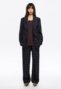 Garnitur w kratę: marynarka i spodnie w kolorach granatowym, brązowym i pomarańczowym. Warstwowo zestawiony z ciemnym topem na ramiączkach z koronkowym wykończeniem. Miękki, dopasowany krój.