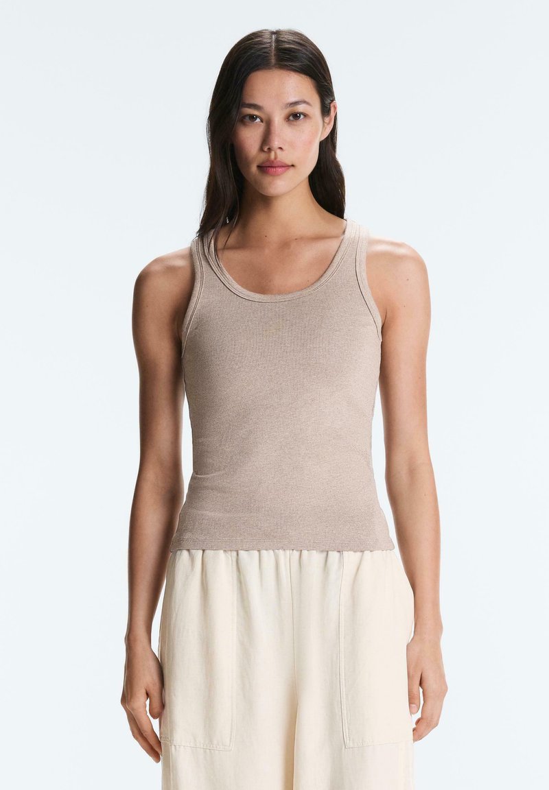 Jeune femme aux longs cheveux foncés portant un débardeur côtelé beige et un pantalon ample couleur crème, debout sur un fond clair uni.