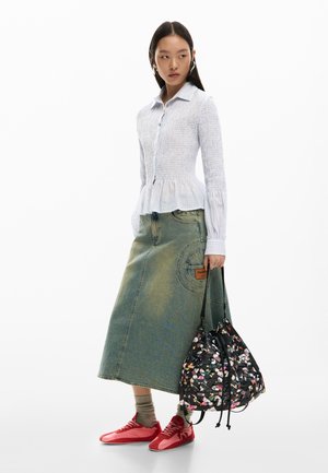 Žena v svetlomodrej blúzke so záhybmi, rifľovej midi sukni, sivých ponožkách, červených teniskách a držiaca čiernu kvetinovú tašku v tvare vedra.