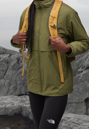 Persoon draagt een olijfgroene jas en zwarte The North Face legging, houdt gele rugzakriemen vast en staat op rotsachtig terrein.